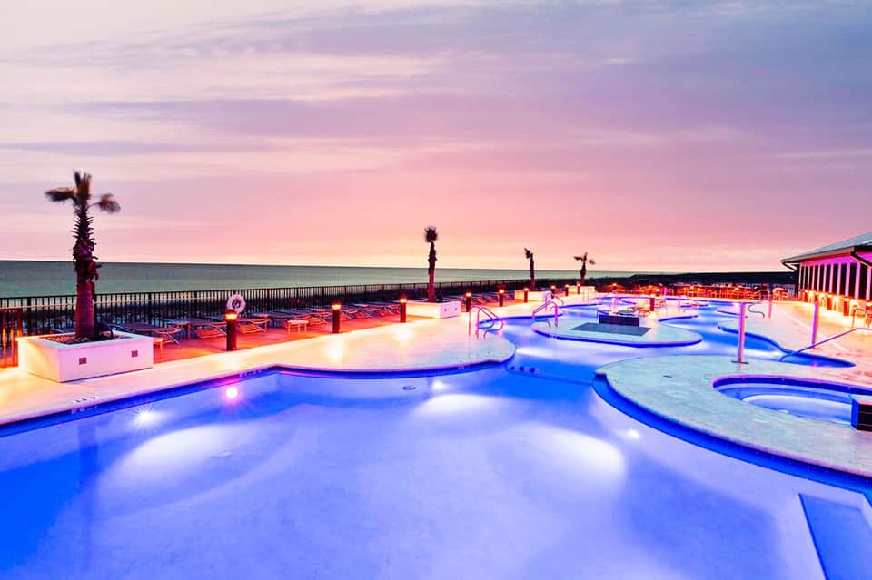 Illuminated resort pool complex at dusk with palm trees and ocean view
