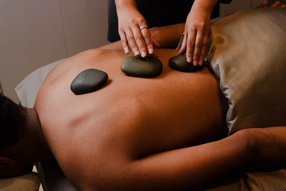 Spa therapist performing hot stone massage on client's back with smooth black stones