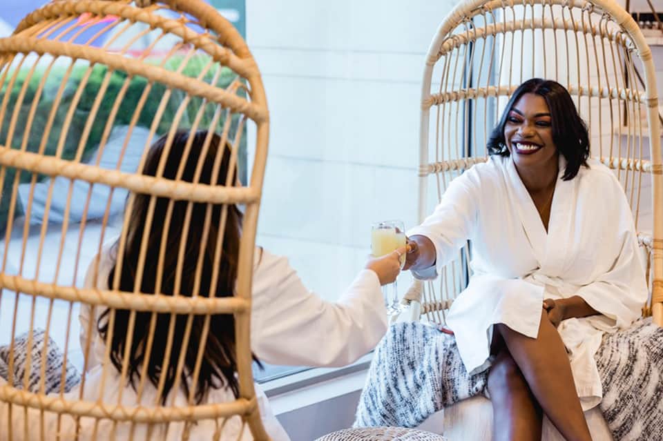 Woman in white robe sitting in hanging rattan chair holding beverage in spa relaxation area