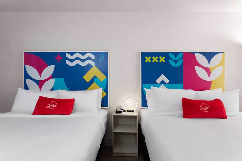 Modern hotel room with two queen beds, colorful geometric headboard artwork, and red accent pillows