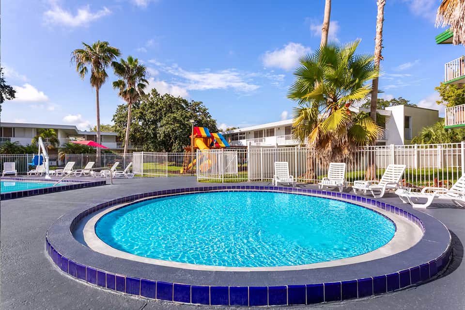 Circular resort pool with blue water, white loungers, palm trees, and colorful waterslide in fenced area