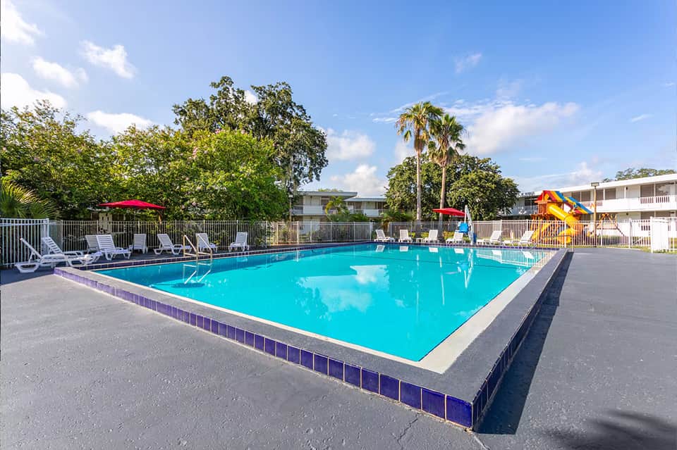 Bright blue rectangular swimming pool with lounge chairs, palm trees, and yellow waterslide