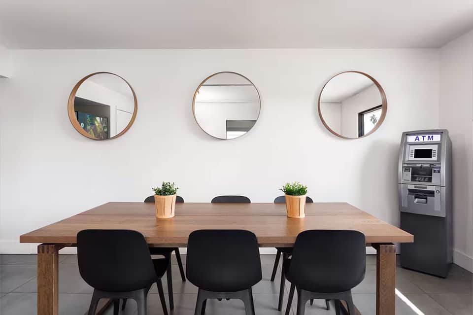 Minimalist meeting room with wooden table, black chairs, three round mirrors, and ATM machine on white walls.