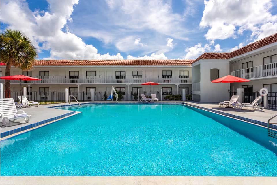 Outdoor pool surrounded by white hotel buildings, lounge chairs, and red umbrellas under clear sky