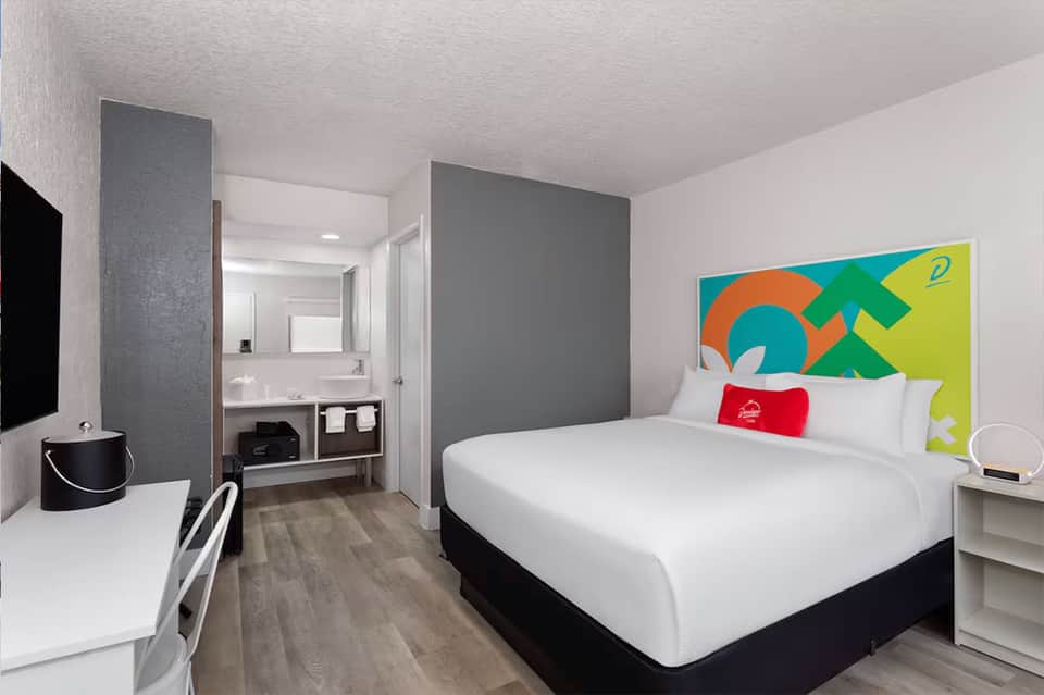 Modern hotel room with queen bed, ensuite bathroom, desk, and colorful abstract headboard art