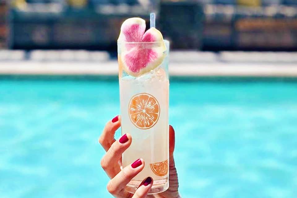 Tall clear cocktail with citrus slices and pink flower garnish held over pool background