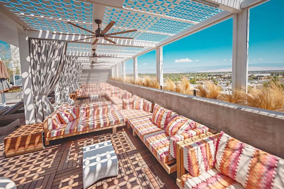 Rooftop lounge with turquoise lattice ceiling, striped cushioned seating, decorative grasses, and panoramic city views