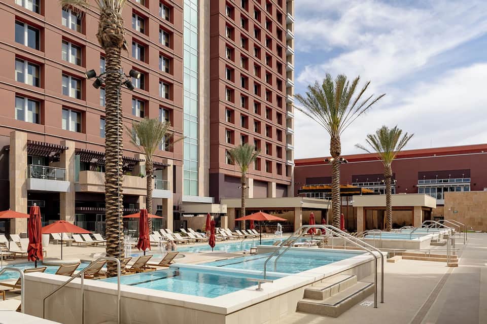Urban resort pool area with red umbrellas, lounge chairs, tall hotel buildings, and tropical landscaping