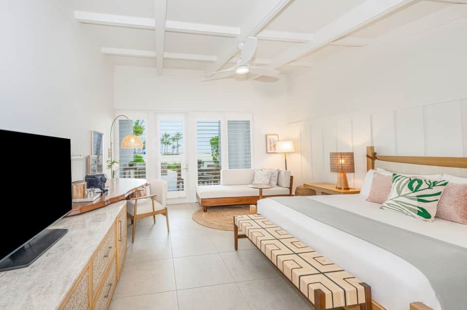 Bright hotel suite with queen bed, sitting area, TV, and ocean-view balcony doors