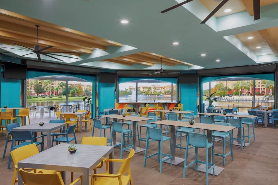 Colorful casual dining area with turquoise and yellow chairs, high-top tables, and waterfront windows