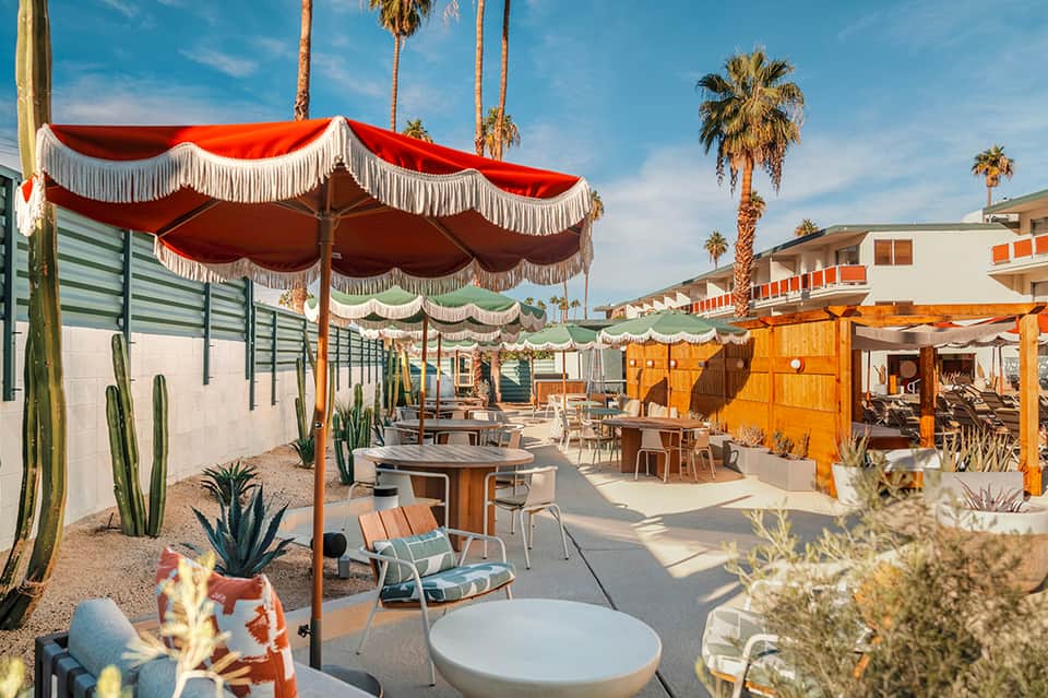 Colorful desert resort courtyard with red and green umbrellas, palm trees, and lounge seating