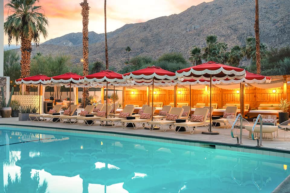Resort pool at sunset with red-and-white striped cabanas, lounge chairs, palm trees, and desert mountain backdrop