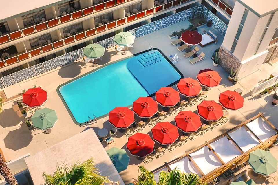 Aerial view of resort pool with red umbrellas, lounge chairs, and multi-story building backdrop