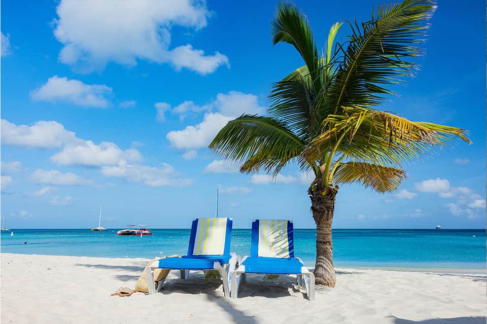 Two blue and white striped beach lounge chairs under palm tree on white sand beach