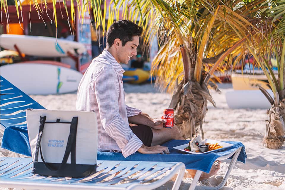 Man relaxing on beach under palm tree with Wyndham Rewards cooler and refreshments