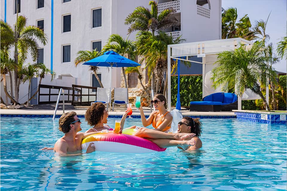 Family enjoying drinks in resort pool with white building and palm trees in background