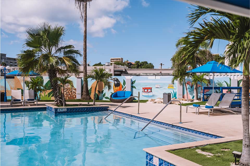 Resort pool area with palm trees, blue umbrellas, lounge chairs, and beachfront mural backdrop