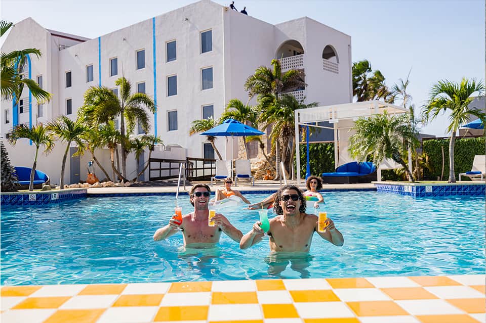 Outdoor resort pool with white building facade, blue umbrellas, palm trees, and guests enjoying tropical pool atmosphere