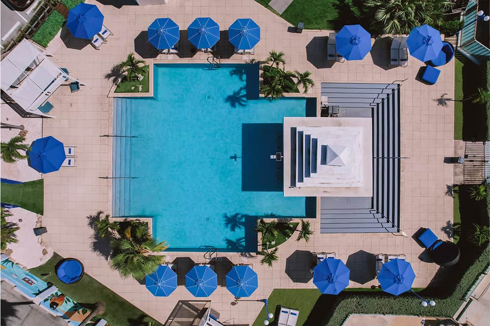 Aerial view of resort pool surrounded by blue umbrellas, palm trees, and manicured lawns