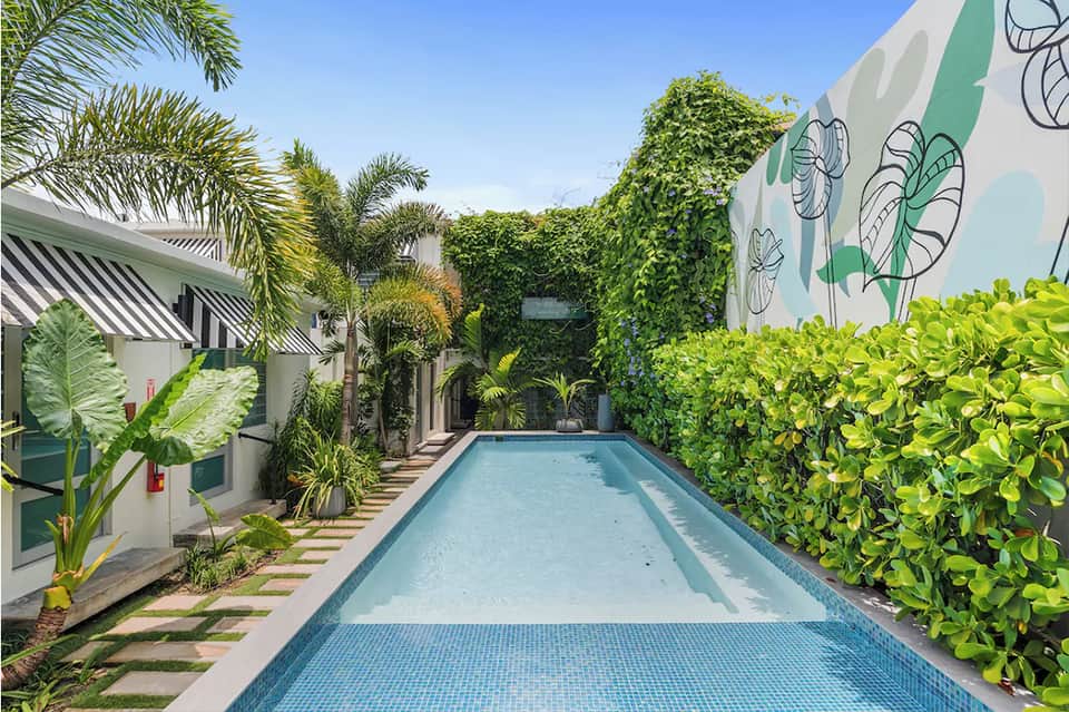 Narrow lap pool surrounded by lush greenery, palm trees, and contemporary white buildings with striped awning