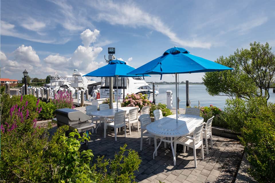 Waterfront dining patio with blue umbrellas, white tables and chairs, boats, and flowering plants