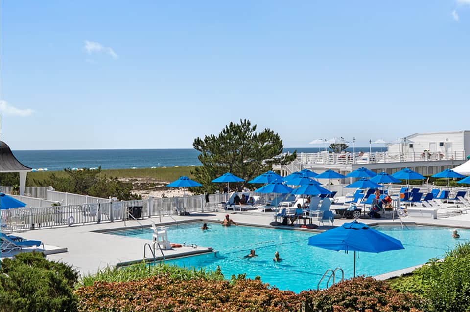 Resort pool area with bright blue umbrellas, loungers, ocean view, and white buildings in background