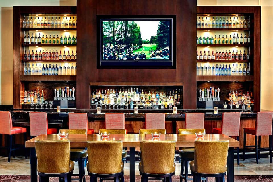 Upscale bar lounge with illuminated bottle displays, dark wood backdrop, and TV screen with gold-accented seating.