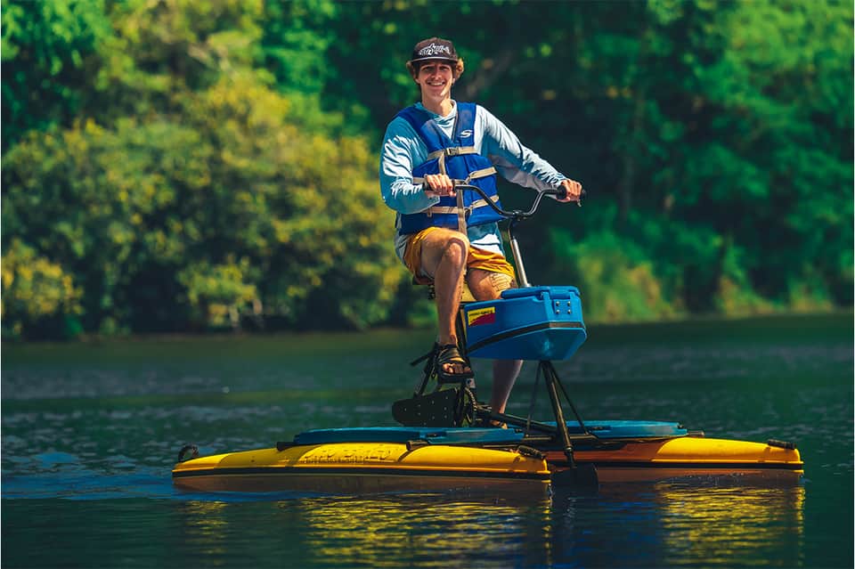 Person on yellow and blue recreational water vessel on river surrounded by green trees