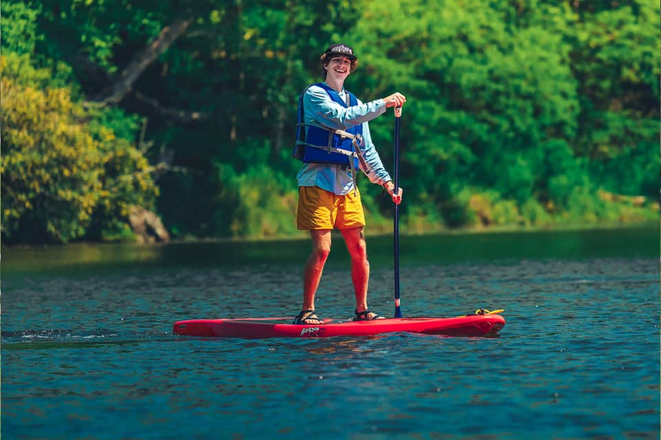 Person stand-up paddleboarding on calm river surrounded by green trees and foliage