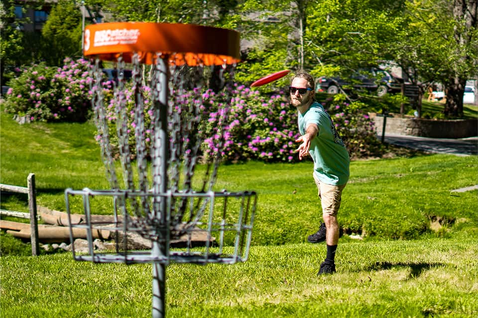 Man throwing disc golf disc on grassy course with purple flowers and orange basket