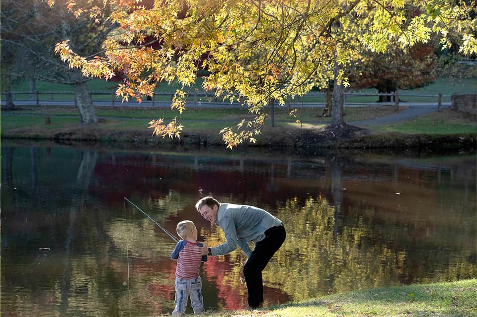 Adult and child fishing by a peaceful pond surrounded by autumn foliage and trees