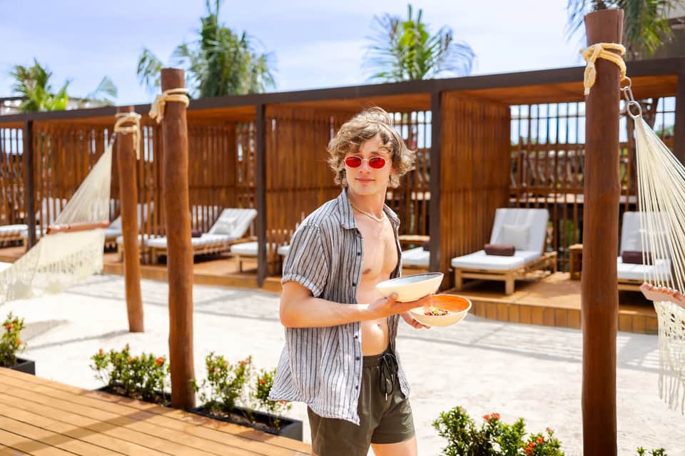 Beachfront dining area with wooden pergolas, palm trees, and server holding food plates