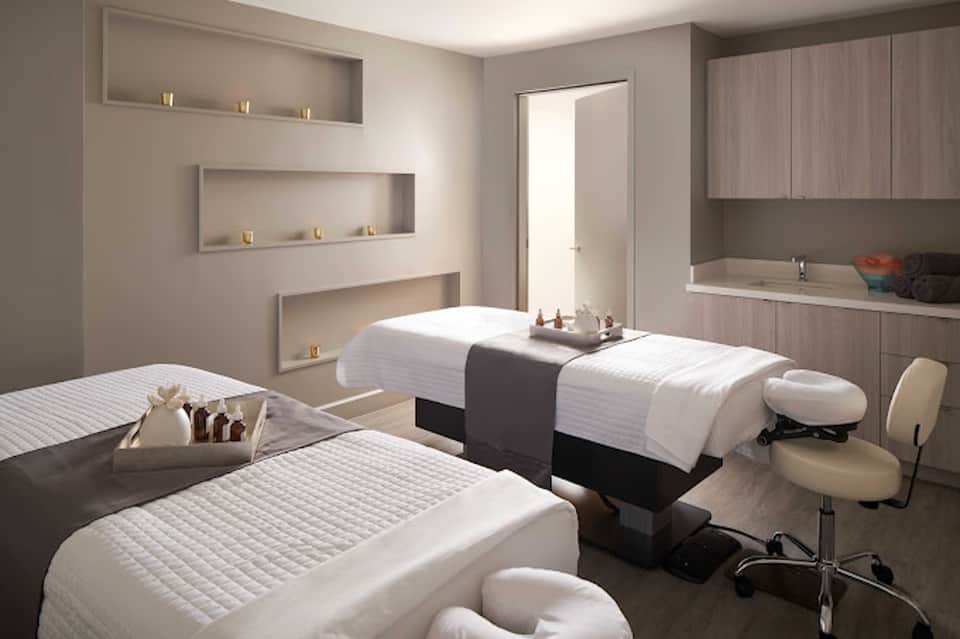 Modern spa treatment room with massage beds, recessed wall shelving, built-in bathtub, and neutral tones