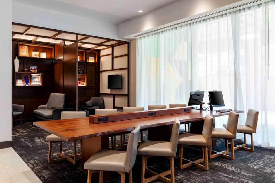 Modern hotel conference room with wooden table, beige chairs, dark wood cabinetry with shelving, and floor-to-ceiling windows