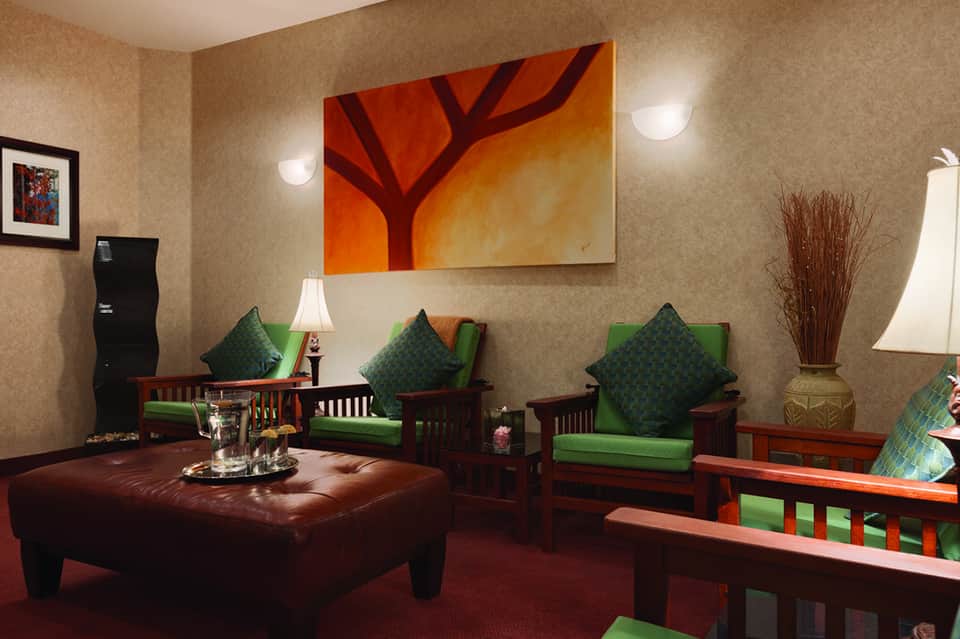 Lounge with tan textured wallpaper, red leather ottoman, green upholstered chairs, and abstract orange tree artwork