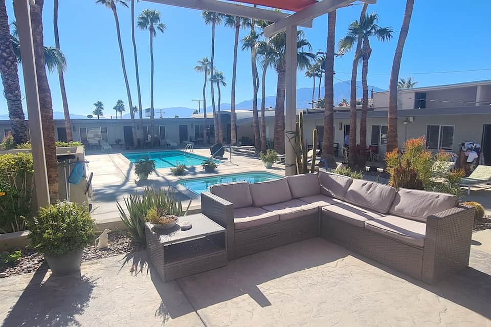 Desert resort lounge area with L-shaped seating, two pools, and palm trees under pergola shade