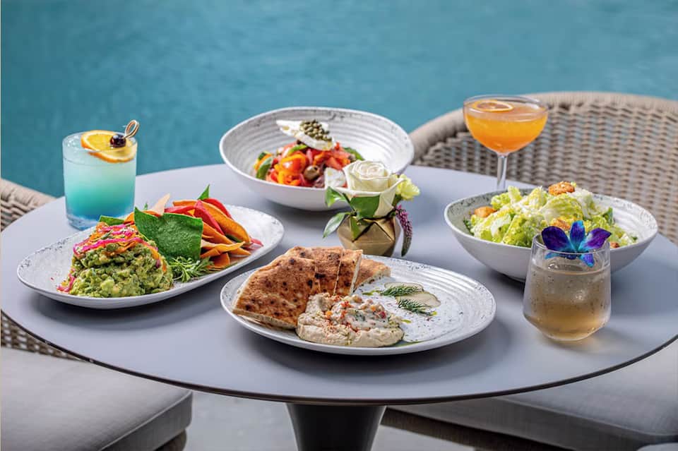 Poolside dining table with gourmet plated dishes, cocktails, and colorful edible flowers on white dinnerware