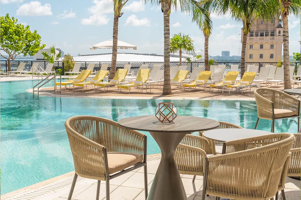 Luxury poolside lounge with palm trees, yellow chairs, and waterfront bridge views
