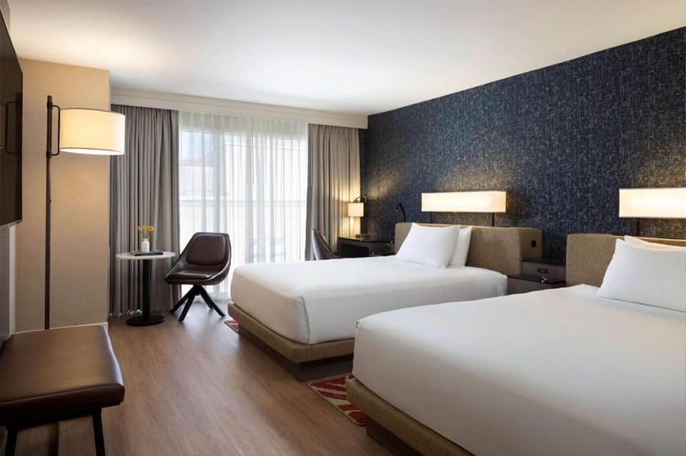Contemporary hotel room with two beds, dark blue accent wall, desk, chair, and floor-to-ceiling windows