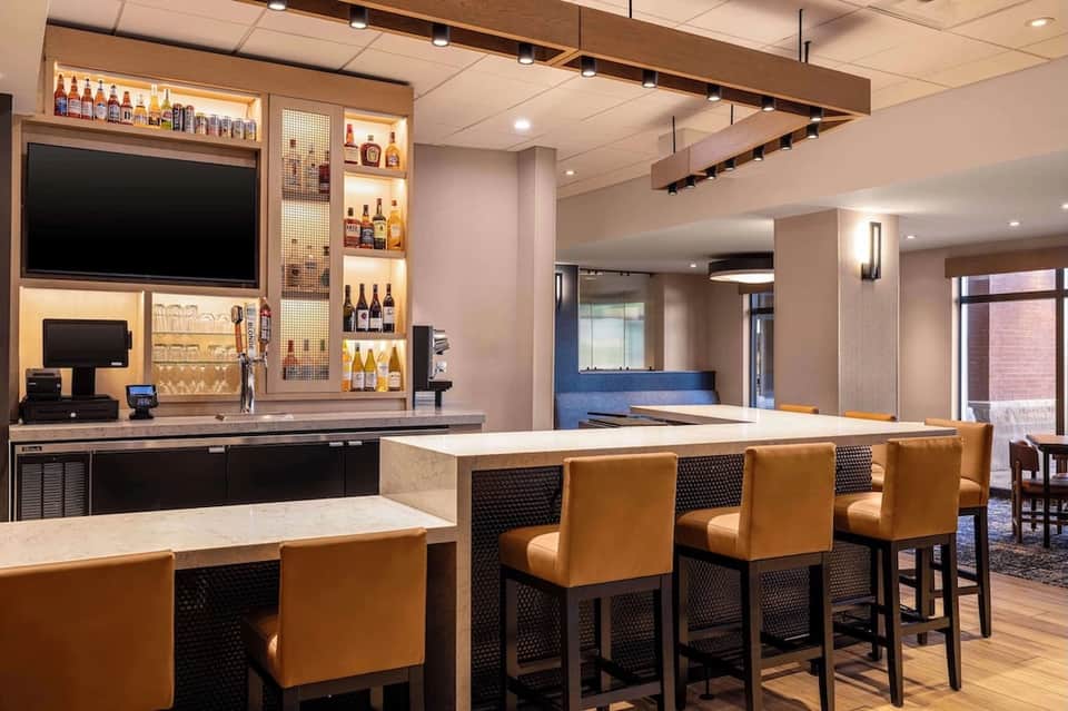 Modern hotel lounge with tan leather bar seating, illuminated liquor shelves, TV, and polished concrete flooring