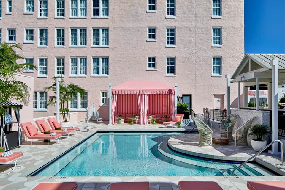 Hotel courtyard with rectangular pool, lounge chairs, pink cabanas, and modern building