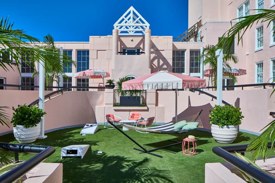 Retro hotel courtyard with pink umbrellas, hammocks, white pavilion, and tropical plants