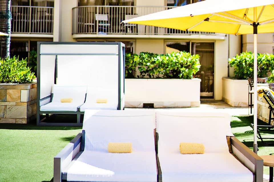 Modern poolside lounge with white sunbeds, yellow umbrella, manicured lawn, and building in background