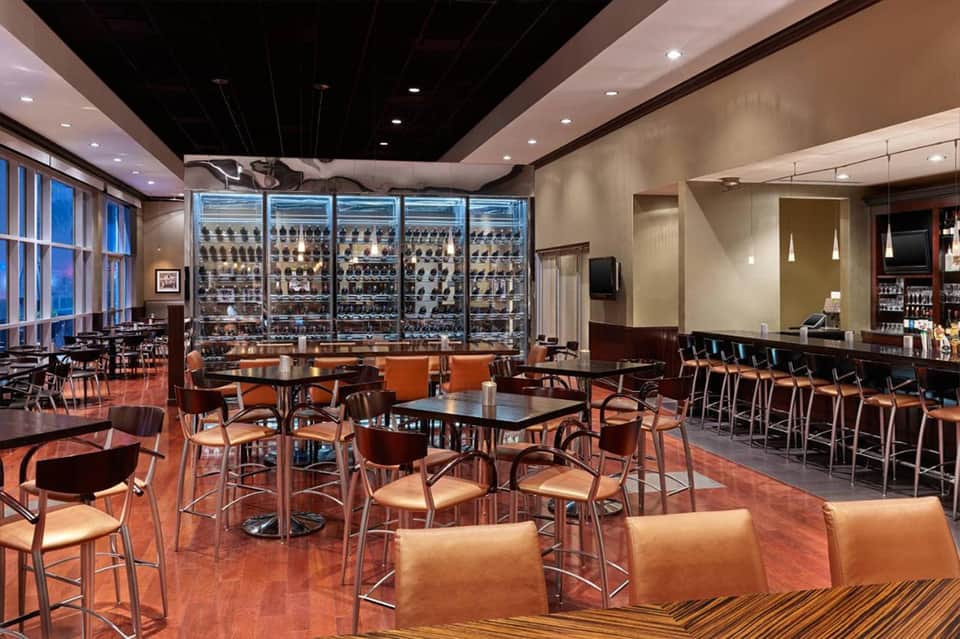 Modern lounge with wine display fridge, orange seating, and floor-to-ceiling windows overlooking city