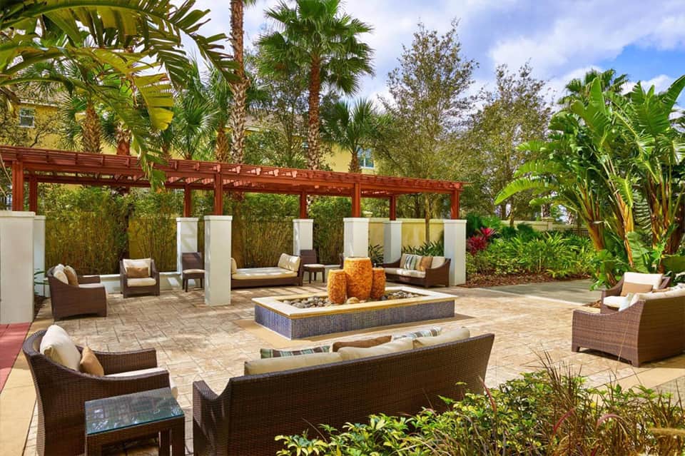 Outdoor courtyard lounge area with rattan furniture, fire pit, pergola, and tropical landscaping