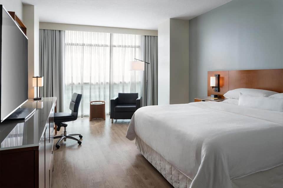 Modern hotel bedroom with king bed, work desk, armchair, and floor-to-ceiling windows