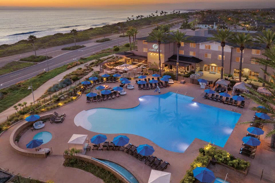 Aerial view of beachfront resort pool complex lit at dusk with blue umbrellas