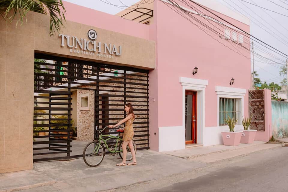 Pink boutique hostel exterior with black slatted gate and woman on bicycle