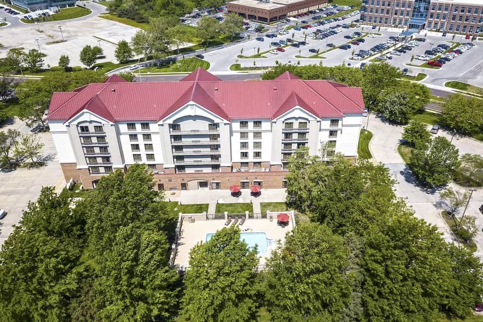 Aerial view of multi-story hotel with red roof, white walls, surrounding parking lot and green trees