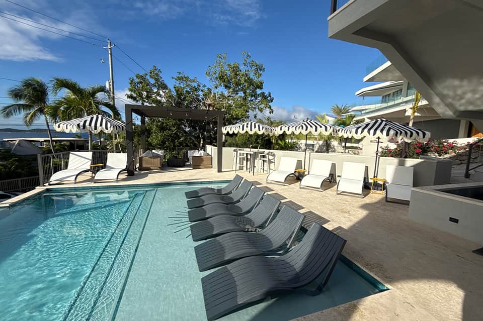 Outdoor pool area with lounge chairs, striped umbrellas, tropical landscaping, and waterfront views